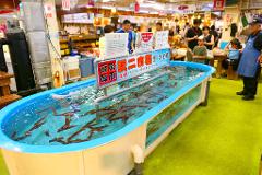 This photo captures a bustling Japanese market scene with a tank full of live squid, where customers are cautioned to avoid touching the water due to potential splashing.