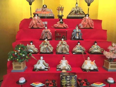 A tiered display of Hina dolls in traditional Japanese attire, arranged on a red fabric backdrop with a yellow wall behind, accompanied by miniature accessories and potted plants.
