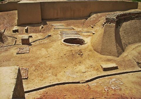 This image shows an archeological excavation site with a brick-lined well, various brick structures, and earthen walls, all set in a sandy, arid environment.