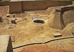 This image shows an archeological excavation site with a brick-lined well, various brick structures, and earthen walls, all set in a sandy, arid environment.
