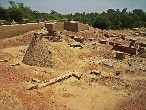 An archaeological site reveals ancient structures, including a conical mound with brickwork, amidst a sandy landscape with scattered brick fragments and ruins.
