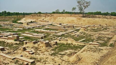 The image shows the ruins of an ancient city, with exposed brick foundations and walls, hinting at a once-thriving civilization, surrounded by earth and sparse vegetation.