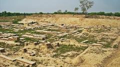 The image shows the ruins of an ancient city, with exposed brick foundations and walls, hinting at a once-thriving civilization, surrounded by earth and sparse vegetation.