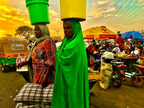 Two women balance buckets on their heads at a bustling outdoor market in Africa, with vibrant colors and busy scenes.