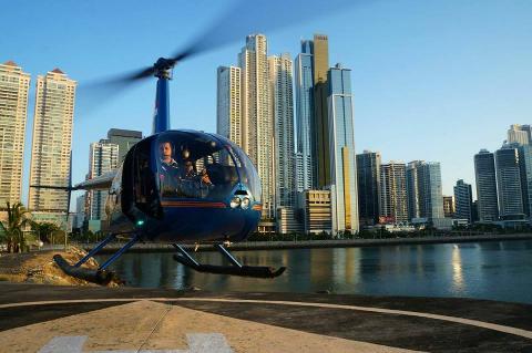 A blue helicopter with two people in the cockpit prepares to take off in front of a skyline of tall buildings and a body of water.
