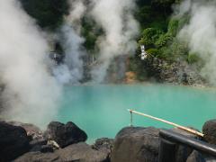 A steaming turquoise pool is surrounded by rocks, green foliage, and plumes of vapor rising into the air at a Japanese hot spring.