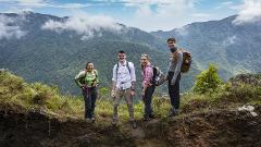 Four hikers stand on a cliff edge, smiling against a backdrop of lush green mountains and a cloudy sky.
