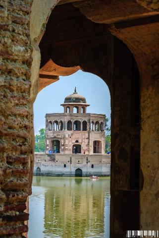 A structure on the water, with an arched entrance in the foreground framing the building and its reflection in the water, with people nearby.
