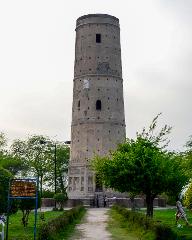 A tall, cylindrical stone tower stands prominently against a pale sky, surrounded by a grassy park with trees and a walking path.