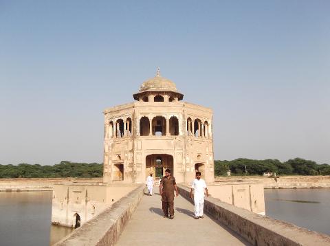 A tan building with a dome sits in the middle of a body of water, with a walkway leading to it and three men walking on it under a clear sky.
