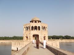 A tan building with a dome sits in the middle of a body of water, with a walkway leading to it and three men walking on it under a clear sky.