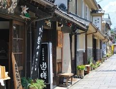 これは、伝統的な商店、日本語の文字が書かれた看板、建物の外にある小さな椅子がある日本の街の風景です。