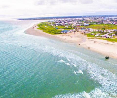 An aerial view captures the serene meeting of a coastline village with the ocean, as waves gently meet the sandy shore under a cloudy sky.