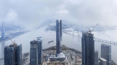 An aerial view of a modern cityscape featuring skyscrapers under construction near a river with bridges, all partially obscured by fog.