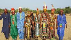 A group of men in traditional African attire, with painted faces and intricate beaded garments, stand holding hands in a cultural celebration or gathering.