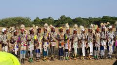 A line of elaborately decorated Wodaabe men with facial paint, headwear, and beaded necklaces, holding staffs in an arid landscape, showcasing their rich cultural attire.