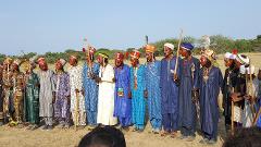 A group of men in colorful traditional attire and face paint stand in a line in a grassy field under a clear sky.