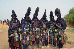 A group of six elaborately dressed men with dark robes and headwear stand in a row, adorned with colorful accessories, in a cultural celebration.