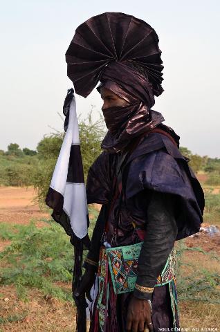 A Tuareg man in Niger, wearing a traditional indigo robe and a turban, holds a black and white flag, standing in a dry landscape with sparse vegetation.