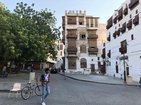 A man walks by a bike near a tree and a chair on a cobblestone street in Al-Balad, Jeddah, Saudi Arabia, with historic buildings with wooden balconies.