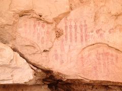 This image shows ancient rock paintings of human-like figures in shades of red and white on a tan sandstone wall, likely found in a cave or sheltered area.