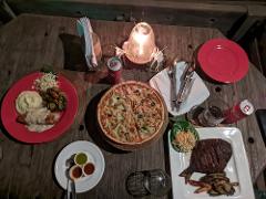 A romantic outdoor dinner featuring pizza, steak, mashed potatoes, and drinks is served on a rustic wooden table under soft candlelight.