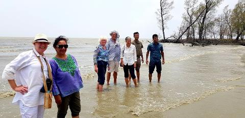 A group of seven people are standing in shallow water on a beach, with trees and cloudy skies in the background.