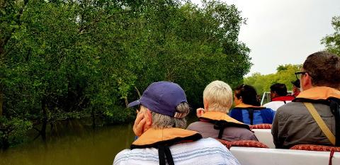 A group of tourists wearing life jackets are on a boat, likely exploring the waterways of Dhaka or the Sundarbans, surrounded by dense mangrove vegetation.