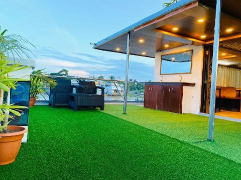 A rooftop deck with artificial grass features seating, potted plants, and a sheltered area with a sink and mirror, overlooking a river scene.