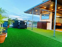 A rooftop deck with artificial grass features seating, potted plants, and a sheltered area with a sink and mirror, overlooking a river scene.