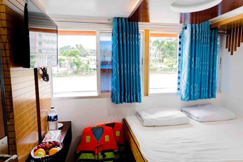 A cozy cabin features a double bed with white linens, flanked by windows dressed in blue curtains, with life vests tucked beside the bed, and a TV, water bottle, and fruit bowl on a small table.