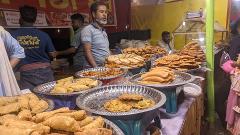 3-Day Dhaka Heritage & Culture Tour — Old Palaces, Lost Cities & Zamindari Mansions A stall displays a variety of traditional Bengali fried snacks and sweets, with people browsing and purchasing them at a cultural event.