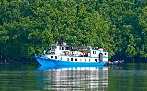 Bangladesh in 7 Days: Dhaka, Sreemongol & the Sundarbans A blue and white boat floats on calm water in front of a dense green forest.