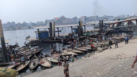 A bustling riverside scene in Dhaka, Bangladesh, features numerous traditional boats docked at a busy ferry terminal.