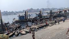 A bustling riverside scene in Dhaka, Bangladesh, features numerous traditional boats docked at a busy ferry terminal.