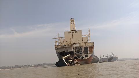 A beached and broken ship rests in the murky waters of Bangladesh, with a distant cityscape visible on the shore under a hazy sky.