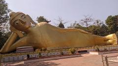 A giant golden statue of a reclining Buddha lies serenely amidst lush greenery under a clear blue sky.