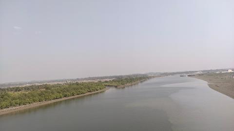 A wide, calm river flows past a lush green treeline and a muddy shore, with a hazy sky overhead.