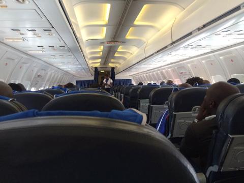 The image shows the interior of an airplane cabin with passengers seated, looking towards the front where a flight attendant stands.