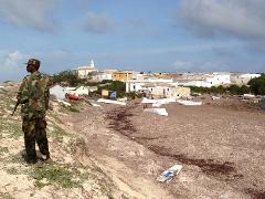 A soldier in camouflage uniform stands guard on a beach with scattered boats near a village under a cloudy sky.