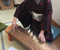 A person wearing a kimono plays a koto, a traditional Japanese stringed instrument, while sitting on a cushion.