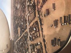 An aerial view from an airplane window shows a dense, arid landscape with buildings and vehicles, possibly in Niger.