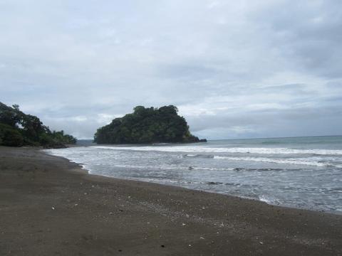 Whale Watching in Nuquí Private Eco Adventure (6 Days / 5 Nights) A secluded, dark sand beach leads to a lush, rocky island and the ocean under a cloudy sky.