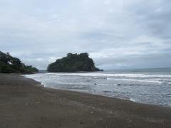 Whale Watching in Nuquí Private Eco Adventure (6 Days / 5 Nights) A secluded, dark sand beach leads to a lush, rocky island and the ocean under a cloudy sky.