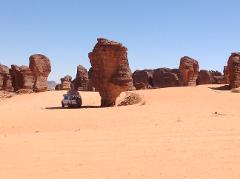 A white SUV is parked in a vast orange desert landscape in Chad, surrounded by unique rock formations under a clear blue sky.