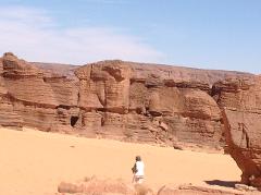 A solitary figure walks in a desert landscape with layered rock formations under a clear, bright sky in Chad.
