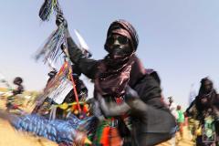 A blurred motion shot captures a Wodaabe man in traditional attire, complete with sunglasses and a headwrap, raising a colorful, tassel-like object during the Bianou & Gerewol Festival in 2019.