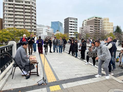 A musician plays the koto on a bridge while a crowd gathers to watch and record, with buildings in the background.
