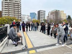 A musician plays the koto on a bridge while a crowd gathers to watch and record, with buildings in the background.