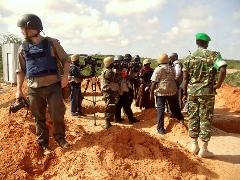 A group of journalists and military personnel stand outdoors on a mound of dirt, with cameras and protective gear, possibly covering a news event.